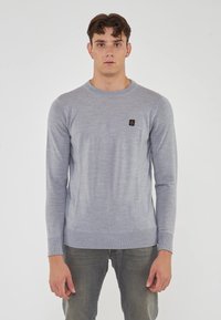 Maglione grigio chiaro a maniche lunghe realizzato in tessuto morbido, con scollo a girocollo, polsini a coste e una piccola toppa con logo scuro sul petto.