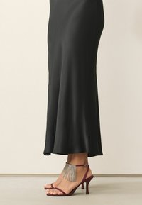 Burgunderfarbene Sandalen mit Riemchen und Absatz, verziert mit einem Strassfransenakzent, kombiniert mit einem einfarbigen schwarzen knöchellangen Kleid aus glattem Stoff.