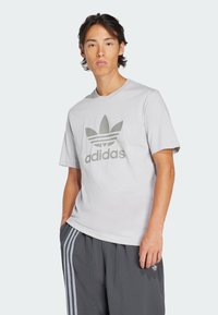 Světlé šedé tričko vyrobené z bavlny, s velkým tónovým logem Adidas a třemi horizontálními pruhy, nošené s texturovanými šedými kalhotami.