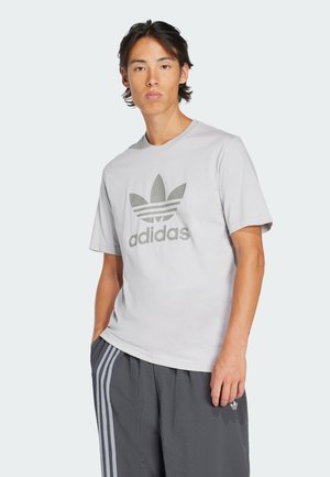Lys grå t-skjorte laget av bomull, med et stort tonet Adidas-logo og tre horisontale striper, brukt sammen med teksturerte grå bukser.