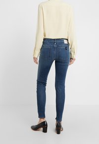 Jeans ajustados de color azul oscuro con un diseño ceñido, que cuentan con bolsillos traseros y una etiqueta en contraste, combinados con una blusa amarilla clara y tacones negros.