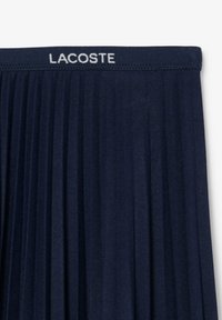 Tamsiai mėlyna klostuota sijonas su elastingu juosmeniu, ant kurio yra baltas "LACOSTE" logotipas. Minkštas audinio paviršius su vertikaliomis klostėmis, užtikrinančiomis judėjimo laisvę.