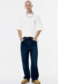 Bílá oversized tričko s malým grafickým designem na kapse, v kombinaci s tmavě modrými volnými džíny a hnědými nazouvacími botami. Viditelný stříbrný řetízek.