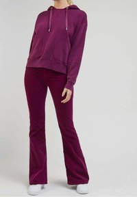 Lila bomullshuvtröja med två framfickor och matchande utsvängda byxor. Materialet verkar mjukt med en jämn textur; avslutad med vita skor.