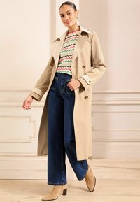 Beige trench coat dengan depan double-breasted, dipasangkan dengan sweter bermotif zigzag berwarna-warni dan celana jins wide-leg biru gelap. Sepatu bot pergelangan kaki berwarna tan.