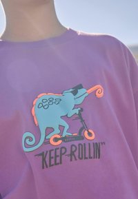 Paarse sweatshirt met een gekleurde kameleon die op een scooter rijdt, met heldere blauwe, oranje en zwarte accenten. De tekst zegt "HOU DOOR."