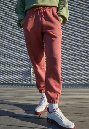 Personne portant un pantalon de jogging couleur rouille, des chaussettes blanches et des baskets blanches avec des semelles rouges, debout sur un sol en bois devant un mur métallique texturé.