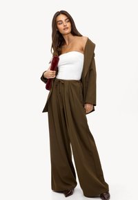 Vrouw in olijfgroene wijde broek, bijpassende blazer van de schouder, witte strapless top, met een rode clutch en donkerpuntige schoenen.