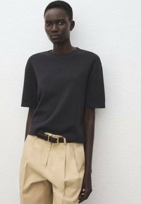 Czarny krótki top z dzianiny z krótkim rękawem zestawiony z beżowymi spodniami w wysokim stanie oraz brązowym paskiem ze złotą klamrą na jasnym tle.