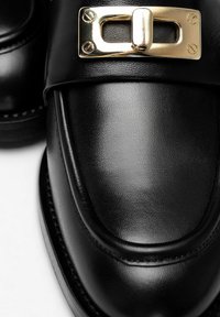 Scarpe di pelle nere con finitura liscia, punta arrotondata e dettaglio della fibbia metallica dorata. Le cuciture aggiungono texture.