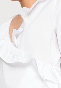 Blusa bianca con colletto arricciato, tessuto texture, lacci regolabili ai lati e silhouette aderente.