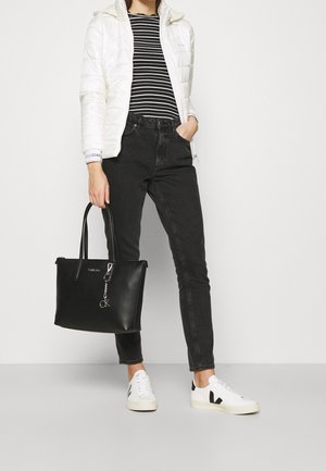 Person iført hvid pufferjakke, sort-hvid stribet bluse, sorte jeans, hvide sneakers, der holder en stor sort læder tote taske med en nøglering.