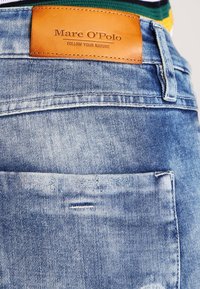 Jeans de mezclilla azul claro con un bolsillo trasero visible, que presenta una etiqueta de cuero marrón que dice "Mare O'Polo" y un tejido texturizado.