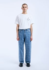 Camiseta blanca de gran tamaño con diseño impreso, combinada con jeans de mezclilla azul que presentan una textura deslavada y un patrón gráfico. El modelo lleva zapatos negros.