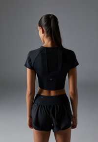 Crni kratki crop top s kratkim rukavima i izrezanom pozadinom, uparen s crnim kratkim hlačama visokog struka. Glatka tekstura, uski kroj.