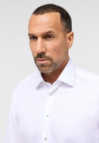 Hvid skjorte med knaplukning og et struktureret mønster, der har en spredt krave og sølvfarvede knapper på stropperne.