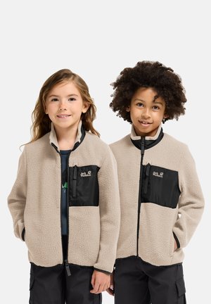 Two smiling children wearing beige fleece jackets with black chest pockets and black pants, standing side by side against a plain background.
