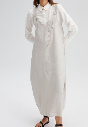 Mujer con vestido midi sin mangas beige con botones delanteros, sobre una camisa blanca de manga larga con cuello, y sandalias negras.