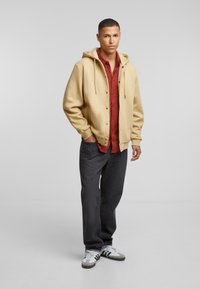 Beige hoodie met knopen aan de voorkant, gedragen over een rood shirt. Gecombineerd met donkergrijze jeans en witte sneakers met grijze accenten.