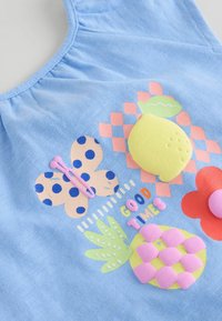 Blaues Oberteil aus Stoff mit Schmetterlings-, Zitronen- und Blumenmotiven in Pink, Grün und Orange, verziert mit strukturierten Akzenten und dem Text "GUTE ZEITEN."