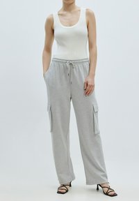 Grijze cargo sweatpants met zijzakken, elastische tailleband en strikdetail, gecombineerd met een witte ribgebreide tanktop en zwarte gekruiste hakken.