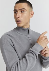 Grauer Reißverschluss-Pullover mit geripptem Textur und hohem Kragen, ausgestattet mit enganliegendem Bündchen. Das Modell gestikuliert mit einer Hand, während es zur Seite schaut.