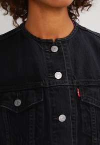 Svart denimjacka med rundad krage, silverfärgade tryckknappar och framfickor. Anmärkningsvärda detaljer inkluderar sömmar och ett litet rött märke.