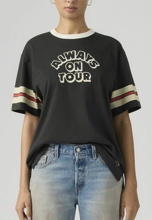 Person iført sort T-shirt med "Always on Tour" tekst og creme og røde striber på ærmerne, kombineret med lyseblå jeans.