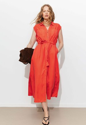 Femme aux cheveux ondulés portant une robe midi orange sans manches boutonnée avec ceinture à la taille, tenant une pochette à motifs foncés, et des sandales noires à lanières.