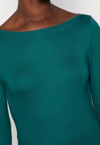 Teal långärmad topp med en bred båtformad halsringning. Tyget är mjukt och lätt, vilket framhäver kroppens konturer utan mönster eller detaljer.