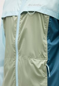 Jachetă Columbia albastru deschis și bej, cu fermoar frontal și buzunar lateral cu fermoar, având sigla pe pieptul superior.