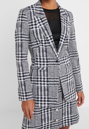 Mujer con blazer y falda a juego de estampado pata de gallo en blanco y negro, sobre una blusa negra con un panel transparente en el escote.