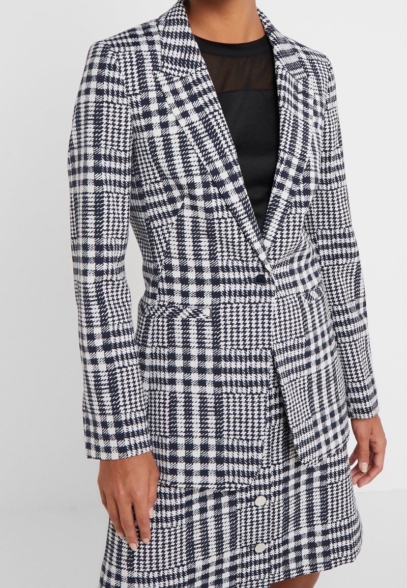 Femme portant un blazer à motif pied-de-poule noir et blanc avec une jupe assortie, sur un haut noir avec un panneau d'encolure en tissu transparent.