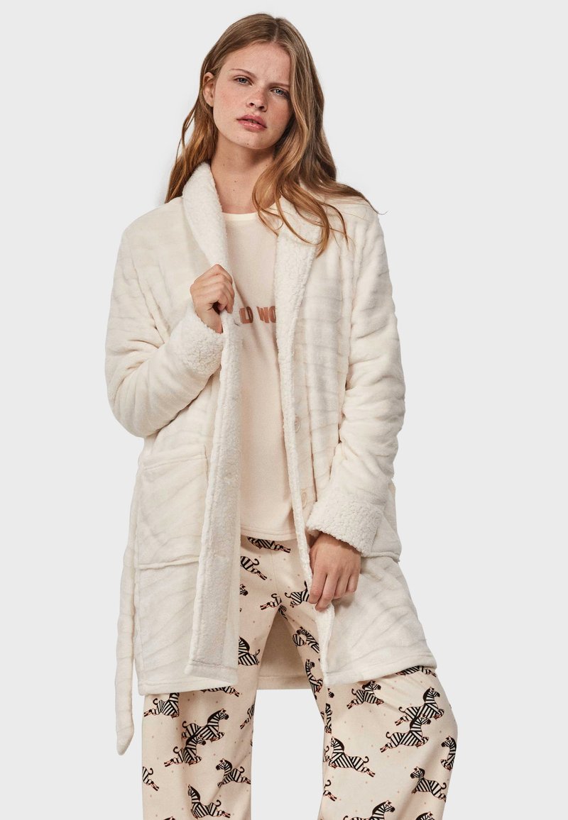 Crèmekleurige pluche badjas met een comfortabele textuur, voorzien van een sjawl-kraag, twee voorzakken en een zelfbindende riem, gedragen over gestreepte pyjamabroeken.
