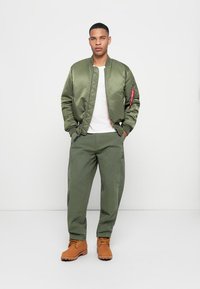 Olive green bomber jacket with a smooth finish, ribbed cuffs, and red zipper accent. Paired with olive cargo pants and tan boots.
