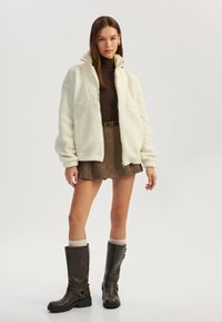 Valkoinen fleece-takki korkealla kauluksella, ruskea poolopaita sen alla, teksturoitu ruskea hame ja polven ylle ulottuvat ruskeat saappaat, joissa on soljet.