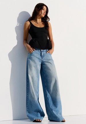 Jonge vrouw met een zwarte mouwloze top en wijde blauwe spijkerbroek, staand met handen in de zakken tegen een witte muur.