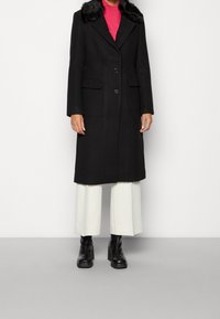 Abrigo negro de lana con cuello de piel sintética, diseño de un solo botón, dos bolsillos frontales, combinado con un suéter rosa y pantalones anchos blancos.