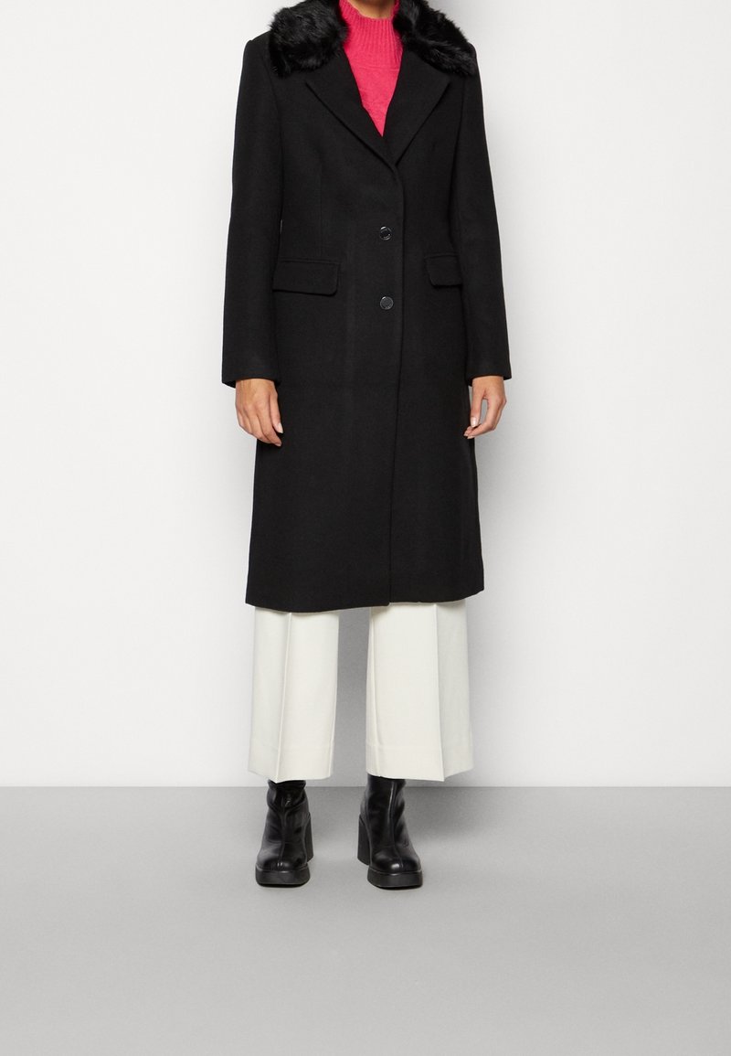 Abrigo negro de lana con cuello de piel sintética, diseño de un solo botón, dos bolsillos frontales, combinado con un suéter rosa y pantalones anchos blancos.
