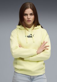 Žltá mikina Puma s kapucňou, predným vreckom, šnúrkami a čiernym logom. Mäkký materiál, uvoľnený strih, manžety na zápästiach a rebrovaný lem.