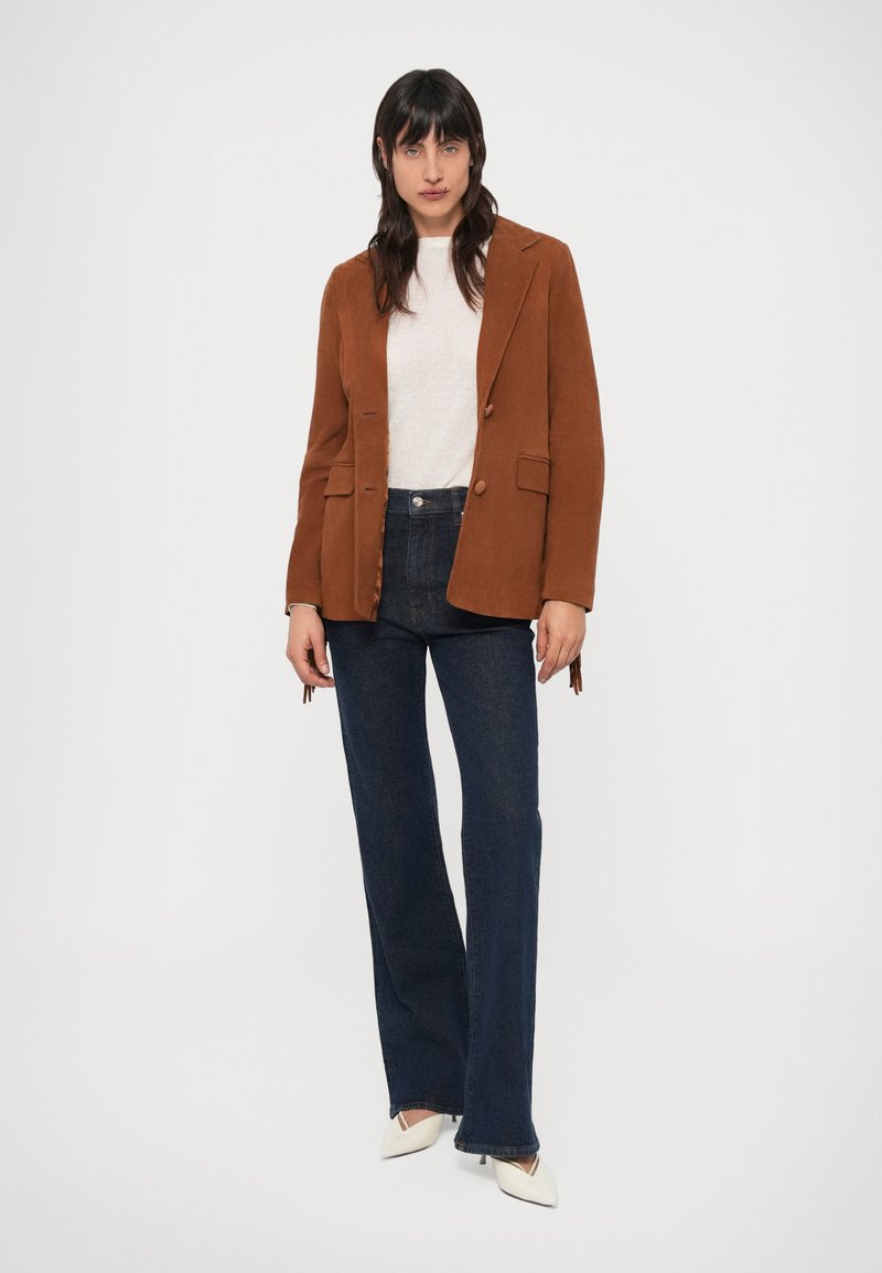 Braunes Cordblazer mit zwei Knöpfen, kombiniert mit einem hellen Pullover, dunklen Schlagjeans und spitzen weißen High Heels. Glatte Textur und maßgeschneiderte Passform.