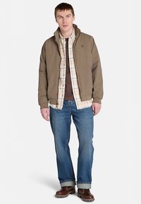 Brun isolerad jacka över en rutmönstrad skjorta, kombinerad med blå jeans och bruna stövlar. Viktiga detaljer inkluderar dragkedja fram och ribbade muddar.