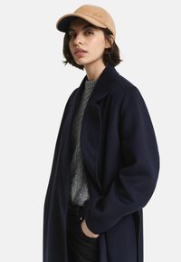Marineblauwe wollen jas met een grote kraag, grijze gebreide trui eronder, en een tan kleurige pet met subtiele branding. Het model heeft een neutrale gezichtsuitdrukking.