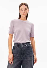Donna con capelli castani lisci che indossa un maglione a maniche corte di colore viola chiaro infilato in jeans a vita alta di un blu scuro scolorito.
