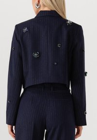 Marineblå stribet cropped blazer med teksturerede blomsterdekorationer, der har en klassisk revers og subtile metalliske accenter.