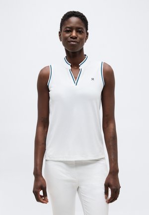 Sieviete ar īsiem matiem, valkā baltu bez piedurknēm polo krekliņu ar zilu apmali un baltas bikses, stāvot pret vienkāršu fonu.