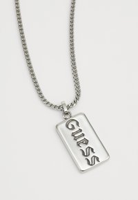 Strieborný náhrdelník s rektangulárnym príveskom so vzorovaným logom "Guess". Hladký povrch a kovový lesk po celom povrchu.