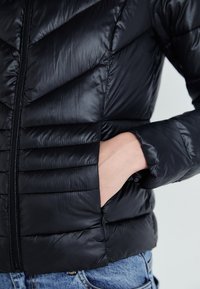 Črna pufferjakna s chevron vzorcem, zadrgo in stranskimi žepi. Mehak, sijoč material; oprijete rukavice in rob.