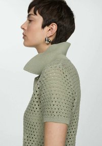 Polo shirt in maglia di colore verde chiaro con colletto alto a costine. Presenta maniche corte e un motivo traforato e texturizzato. Orecchini a cerchio argentati.