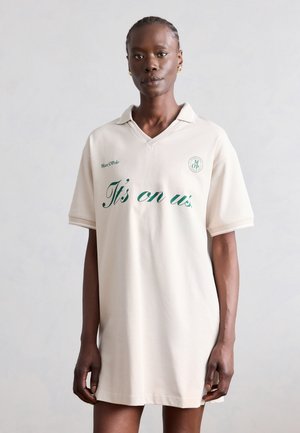 Mustanahaline naine patsidega, kellel on kreemikas polo-kleit rohelise tekstiga "It's on us", seisab ühevärvilise heleda tausta ees.